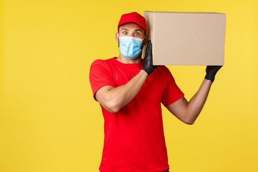 Express delivery during pandemic, covid-19, safe shipping, online shopping concept. Surprised courier in red uniform, medical mask, look excited camera, holding box package on shoulder.
