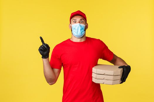 Food delivery, tracking orders, covid-19 and self-quarantine concept. Shocked young courier in red uniform and medical mask, pointing finger upper left corner as deliver pizza to clients house.