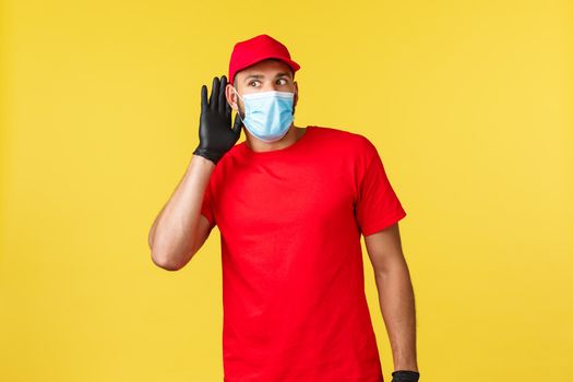 Express delivery during pandemic, covid-19, safe shipping, online shopping concept. Excited and interested courier in red uniform and medical mask, hold hand near ear and eavesdropping conversation.