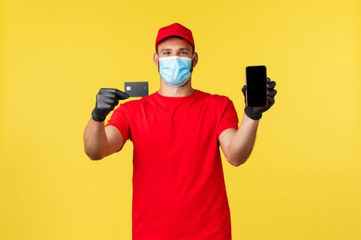 Food delivery, tracking orders, covid-19 and self-quarantine concept. Smiling courier in medical mask and gloves, showing credit card and smartphone, ways of paying for groceries deliver.