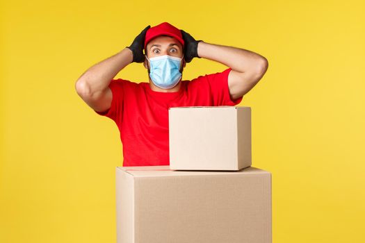 Express delivery during pandemic, covid-19, safe shipping, online shopping concept. Embarrassed and nervous, panicking courier standing near boxes with packages, gasping grab head, wear medical mask.