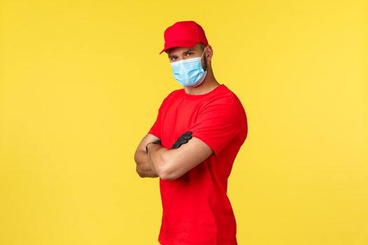 Express delivery during pandemic, covid-19, safe shipping, online shopping concept. Determined handsome courier in red uniform and medical mask, cross arms chest confident look camera.