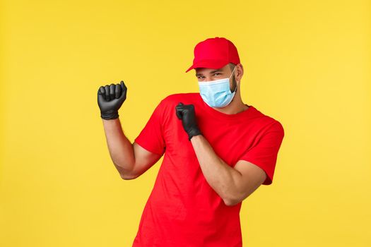 Express delivery during pandemic, covid-19, safe shipping, online shopping concept. Carefree smiling courier in red uniform, medical mask and gloves, dancing, promo of logistic company.