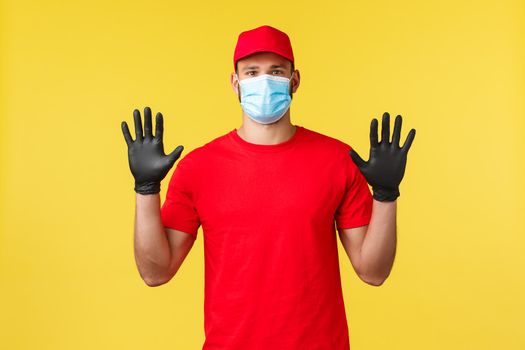 Express delivery during pandemic, covid-19, safe shipping, online shopping concept. Young courier in red uniform, protective medical mask showing clean hands in gloves, employee deliver mail.