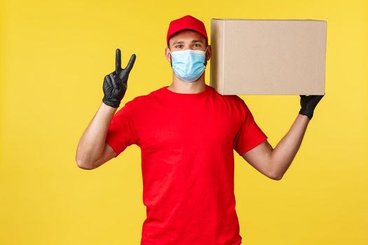 Express delivery during pandemic, covid-19, safe shipping, online shopping concept. Friendly handsome courier in red uniform, medical mask, show peace sign and holding box package.