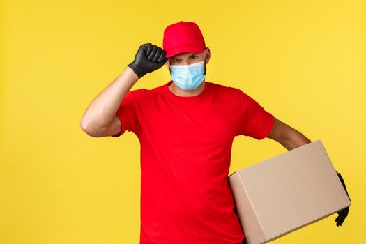 Express delivery during pandemic, covid-19, safe shipping, online shopping concept. Serious determined courier protecting clients package, hold box, saluting with cap, wear medical mask and gloves.