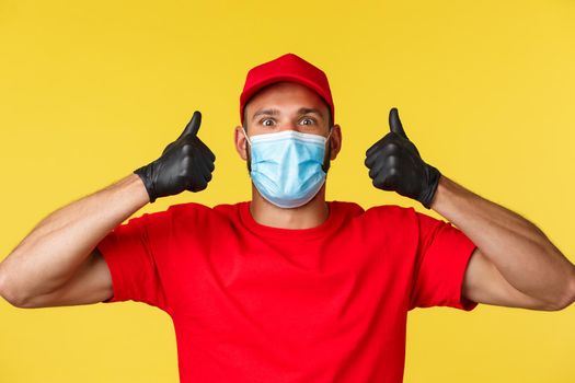 Covid-19, delivery orders, shopping, contactless payment and social distancing concept. Upbeat and excited male courier in red uniform, medical mask, show thumbs-up in approval.