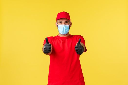 Express delivery during pandemic, covid-19, safe shipping, online shopping concept. Upbeat courier in red uniform, medical mask and gloves, show thumb-up, encourage shop during self-quarantine.