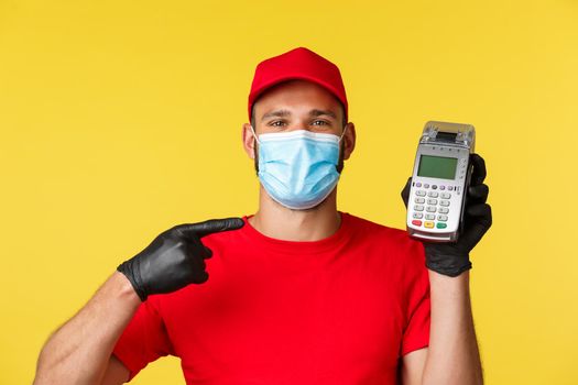 Covid-19, delivery orders, shopping, contactless payment and social distancing concept. Close-up smiling handsome courier in red uniform and medical mask, gloves, pointing finger at payment terminal.