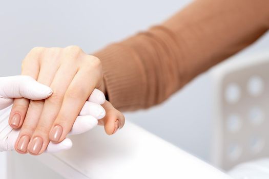 Close up of human hand in protective glove holding female fingers with beige manicure in beauty salon