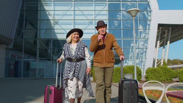 Elderly old husband and wife retirees tourists go to airport terminal for boarding or railway station for traveling. Lovely mature couple grandmother grandfather carrying luggage suitcases on wheels