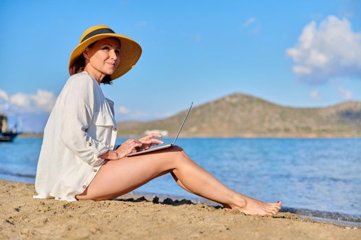 Mature woman in hat using laptop on the beach. Middle-aged female on vacation at the sea, working on a laptop. Remote work, freelance, business, nature, leisure, successful life, 40s people concept