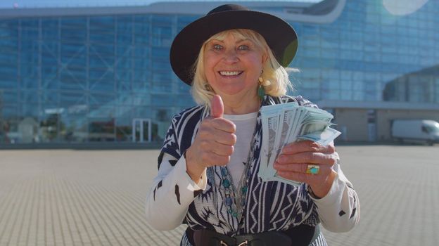 Senior rich pensioner tourist grandmother businesswoman stay near international airport hall or railway station holding cash money in dollar banknotes celebrate success win. Travel, vacation, holidays