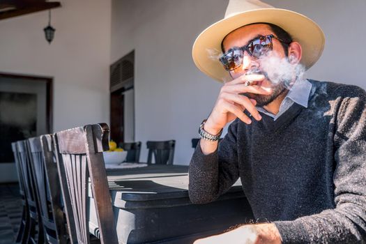 Young Nicaraguan man wearing a sweater hat and sunglasses smoking a cigar in the living room of a colonial-style mansion in Managua city
