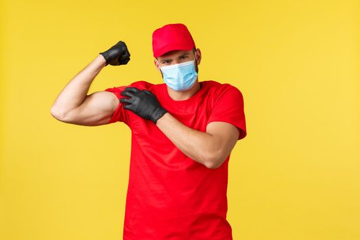 Express delivery during pandemic, covid-19, safe shipping, online shopping concept. Strong courier in red uniform, medical mask and gloves, flex biceps, show-off strong muscles.