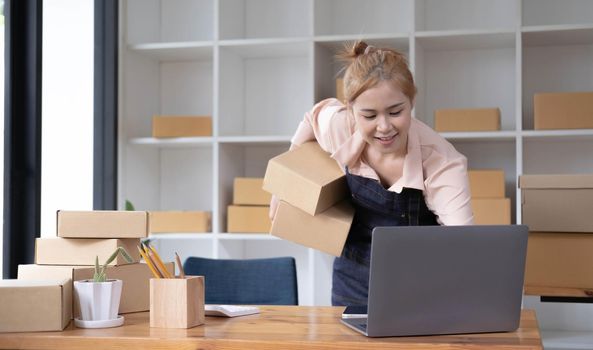 Startup SME small business entrepreneur SME or freelance Asian woman using a laptop with box, Young success Asian woman with her hand lift up, online marketing packaging box and delivery, SME concept..