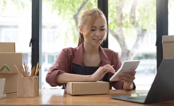 Close up woman sitting in the office full of packages in the background emplaning on a laptop and using a calculator, for online business, SME, finance, e-commerce and delivery concept..