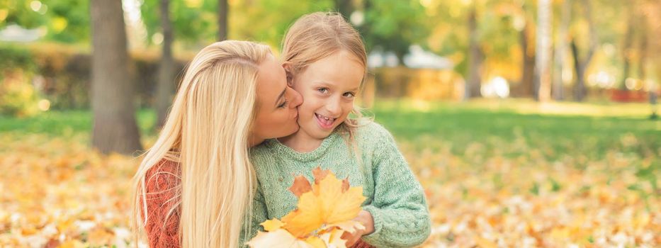 Happy young caucasian mother and little daughter holding autumn yellow leaves sitting and kissing at the park