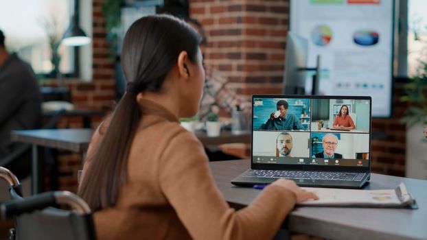 Company employee using online videocall conference on laptop to talk to businesspeople in office. Female worker in wheelchair meeting on remote teleconference call for distant communication.