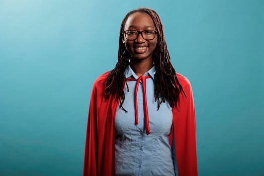 Optimistic looking superhero woman wearing hero costume while smiling heartily at camera on blue background. Studio shot of justice defender wearing mighty cloak while being positive.