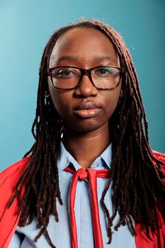 Portrait of brave superhero woman wearing mighty hero red cloak standing on blue background while looking at camera. Strong justice defender with superpower abilities posing for camera.