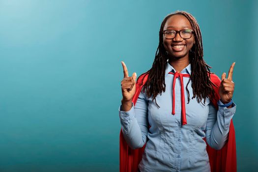 Confident and strong superhero woman wearing red cloak poiting fingers up while standing on blue background. Selfless brave justice defender posing for camera while gesturing fingers up. Studio shot