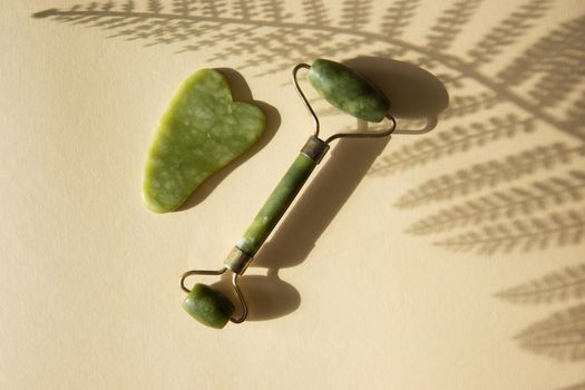 Jade Gua sha scraper and face roller massager on a cork round stand with a monstera leaf. Hard light, shadows, the concept self-care. Facial care. Zero waste. Lifting and toning treatment at home.