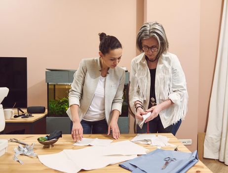 Two pretty seamstresses of different nationalities and ages in a tailoring workshop discussing fabric, sewing pattern and sketch during production process. Colleagues at atelier creating new garment