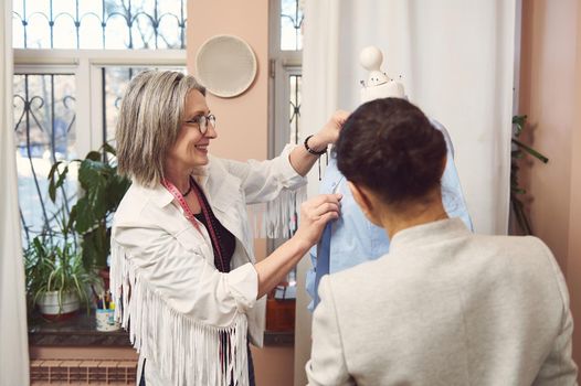 Beautiful mature European female fashion designer and her assistant trying on a new shirt on a mannequin, preparing her new collection of classic clothes for an exhibition in a fashion design studio
