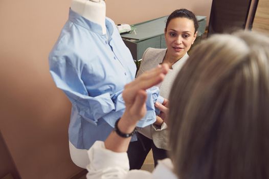 Focus is on charming beautiful middle aged Hispanic fashion designer seamstress tailor working on her new clothing collection on mannequin dummy in tailoring and fashion design workshop
