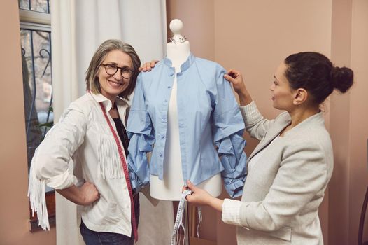 Senior fashion designer smiles posing with a dressmaker model wearing a blue shirt from new collection while her colleague tailor takes measurements and works on improvement details of new garment