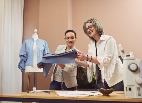 Two charming women- fashion designers team talk about new collection, holding and looking at blue satin cloth, discussing fabrics and sewing patterns for new collection in dressmaker tailoring atelier