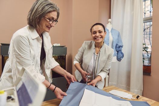 Waist-length portrait of smiling, creative, skilled, inspired mature female fashion designers tailors making clothes patterns on fabric while sewing classic women's shirt in a fashion design workshop