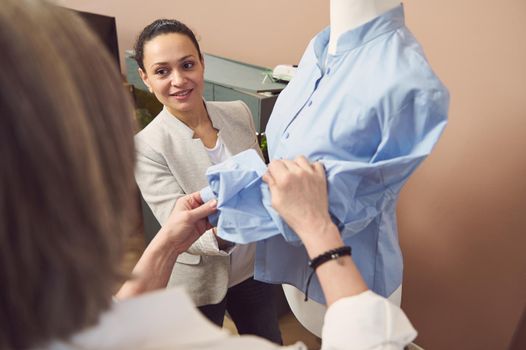 Skilled multi-ethnic team of fashion designers discussing the style and quality of blue shirt fabric on a dressmaker's model in a fashion design and tailoring atelier
