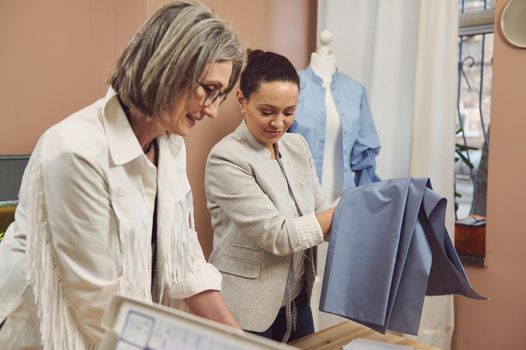 Portrait of a blurred senior fashio designer and middle-aged Hispanic seamstress in a stylish jacket holding blue fabric to create a new collection of women's dress shirts in a fashion design workshop