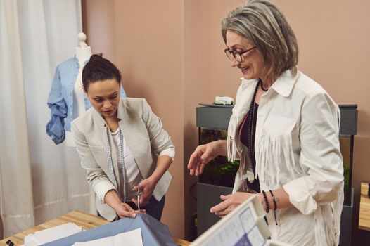Middle aged Hispanic cute woman, skilled tailor cuts a blue fabric pattern under the supervision of a creative senior fashion designer while creating new clothes in the atelier. Seamstress, dressmaker