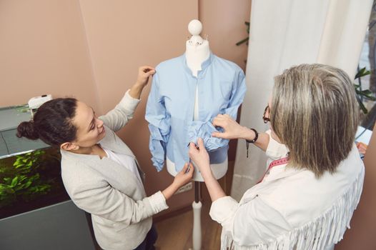 View from above to a team of multi-ethnic and diverse female fashion designers tailors trying on a handmade classical shirt from new collection on mannequin in a fashion design and tailoring atelier