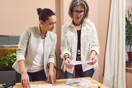 Creative skilled team of two women fashion designers couturiers tailors viewing fashion sketch and discussing sewing pattern while working in modern clothing design and tailoring atelier