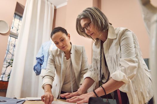 Inspired and skilled team of two diverse women fashion designers seamstresses working with sewing pattern , creating new collection of clothes in a clothing design and tailoring atelier.