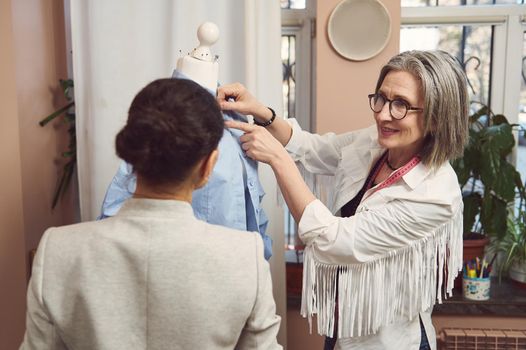 A skilled team of fashion designers tailors try on the new collection of women's dress shirt on a mannequin and work to improve the style and design of the new garments in the tailoring atelier