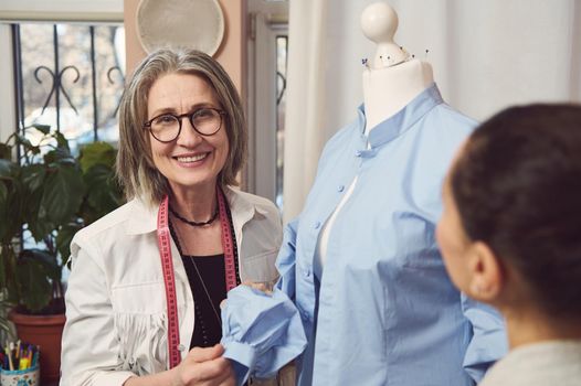 Smiling senior fashion designer try on a new dress shirt on a mannequin while perfecting new collection of clothes in tailoring atelier. Creation of new exclusive handmade clothes. Business and hobby