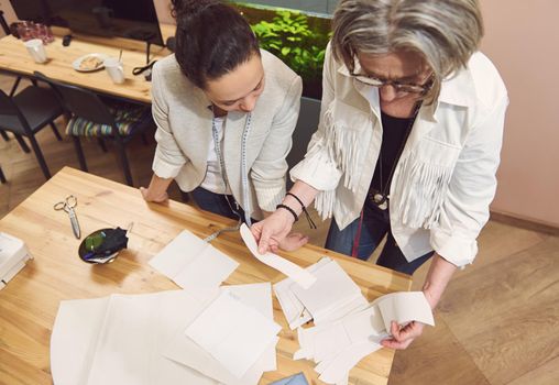 Two stylish women of diverse nationalities and ages discussing sewing patterns while developing a new collection of clothes in a fashion design workshop and tailoring atelier. Tailors, seamstresses
