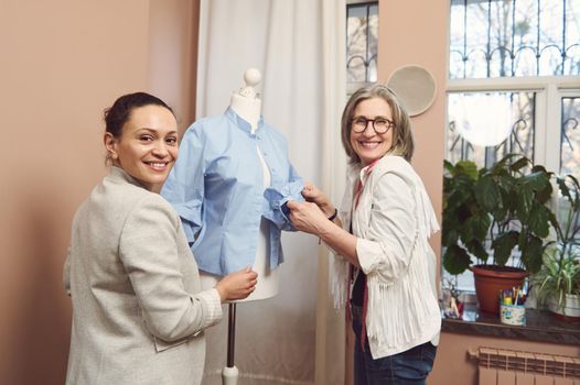 Gorgeous multi-ethnic diverse seamstresses tailors work with a blue shirt on a dressmaker's model, improve details of new collection in a fashion design atelier. Fashion designer business start-up
