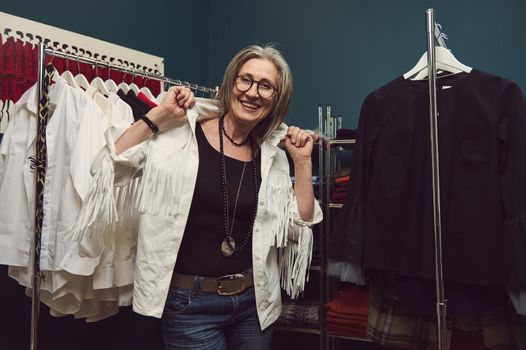 Portrait of happy smiling mature Caucasian woman fashion designer seamstress or tailor posing between hangers with clothes of her new handmade collection in small business fashion or styling workshop.