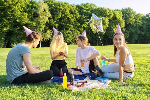 Teenage birthday party picnic on the grass in the park. A group of teenagers having fun, congratulating the girl on her 17th birthday. Age, adolescence, holiday, fun, birthday concept