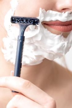 Beautiful young caucasian woman shaving her face by razor on white background. Pretty woman with shaving foam on her face