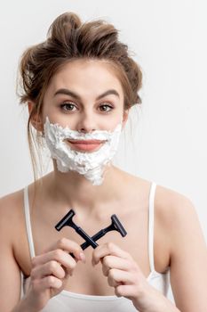 Beautiful young caucasian woman with shaving foam on her face and two razors in her hands on white background