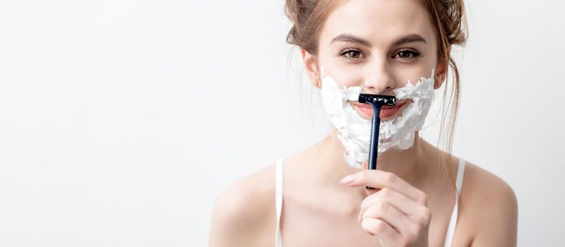 Beautiful young caucasian woman shaving her face by razor on white background. Pretty woman with shaving foam on her face