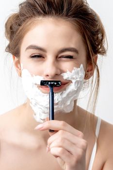 Beautiful young caucasian woman shaving her face by razor on white background. Pretty woman with shaving foam on her face