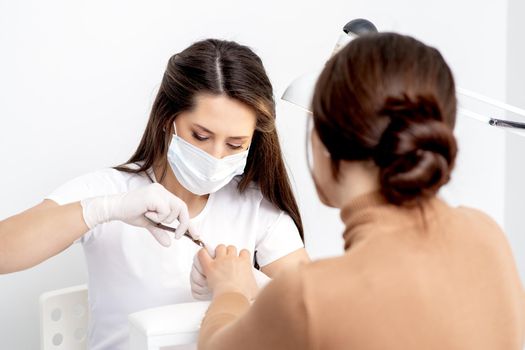 Manicurist in protective mask using manicure cutter to remove cuticle of female nails in manicure salon